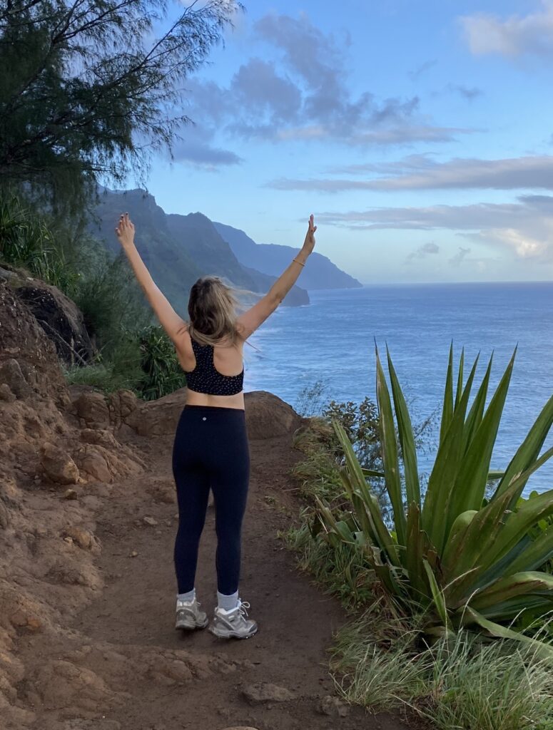 Woman hiking on the Na Pali Coast Trail in Kauai, Hawaii, with arms raised, enjoying the stunning views of the ocean and cliffs.