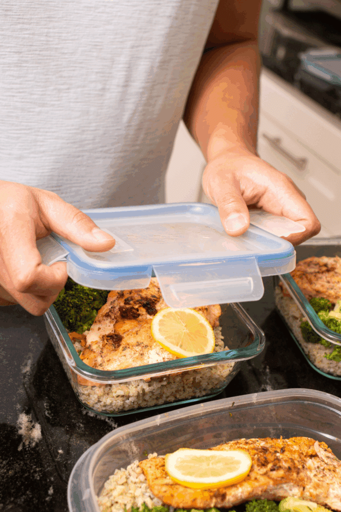 A person placing a lid on a glass meal prep container filled with salmon, lemon, and broccoli on a kitchen counter.