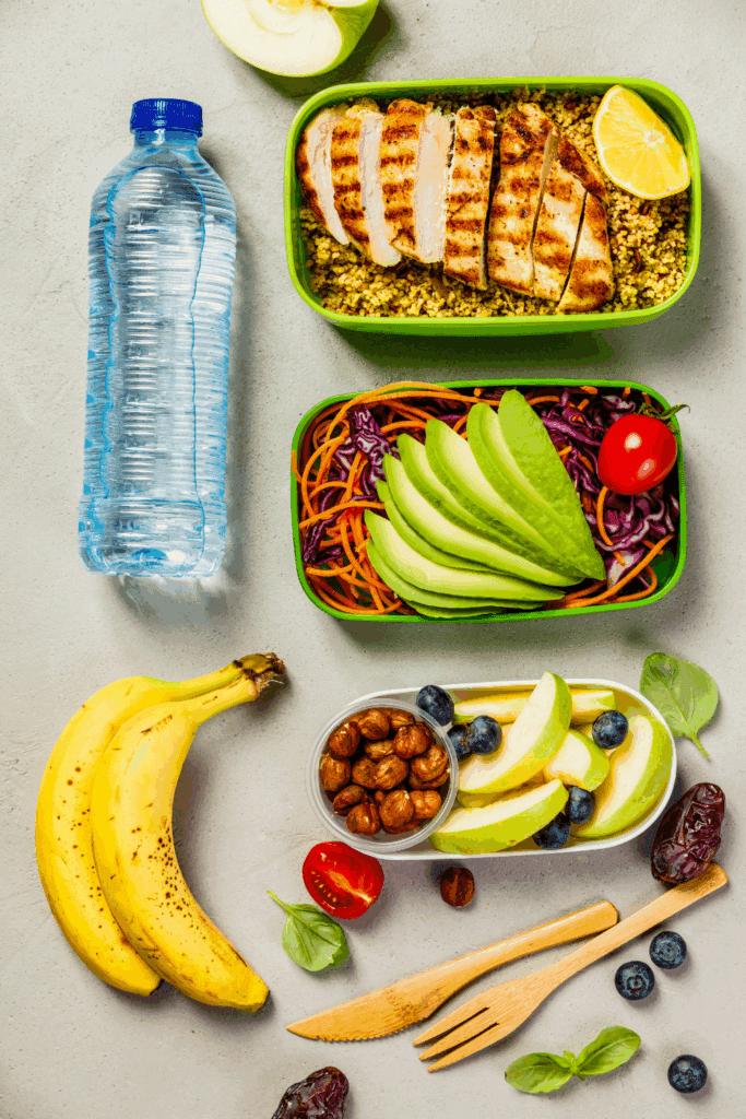 An overhead view of a meal prep scene featuring grilled chicken slices over couscous, a colorful salad with greens and tomatoes, a container of nuts and apple slices, two bananas, a bottle of water, and a wooden fork and knife on a light countertop.