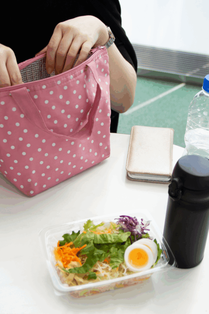 A person is reaching into a pink polka-dotted lunch bag while a container with a fresh salad and a boiled egg is placed on a table alongside a water bottle and a wallet.
