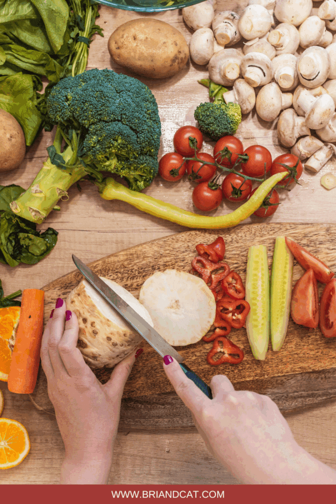 A person slicing root vegetables on a wooden cutting board surrounded by various fresh vegetables including broccoli, tomatoes, mushrooms, and potatoes.