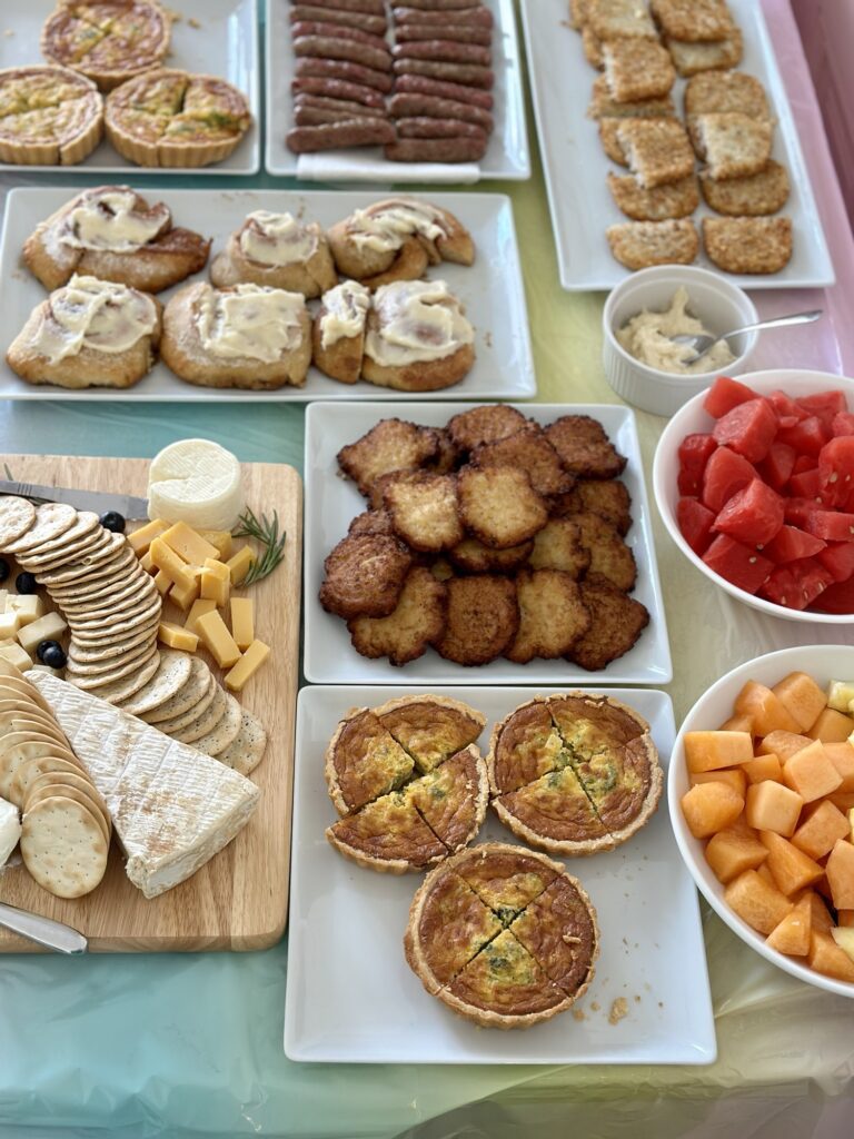 A beautifully arranged brunch spread on a table featuring various dishes, including quiche, cinnamon rolls, sausages, crackers with cheese, and fresh fruits like watermelon and cantaloupe.