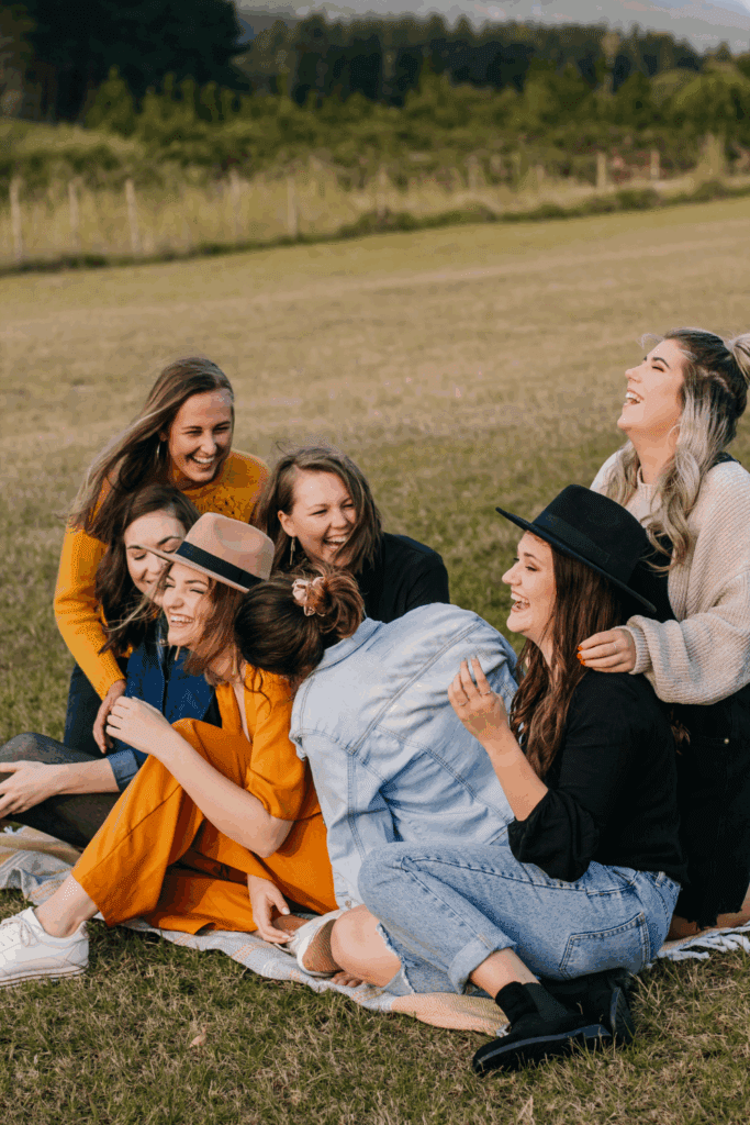 A group of eight women laughing and enjoying each other's company while sitting on a blanket in a grassy field.