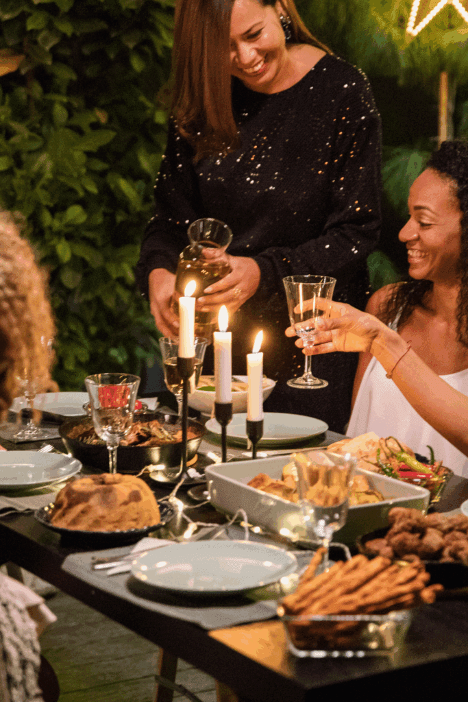A group of friends enjoying a cozy dinner party, with a table set for a meal featuring various dishes, candles, and drinks. One person is pouring a drink while others are engaged in conversation and laughter.