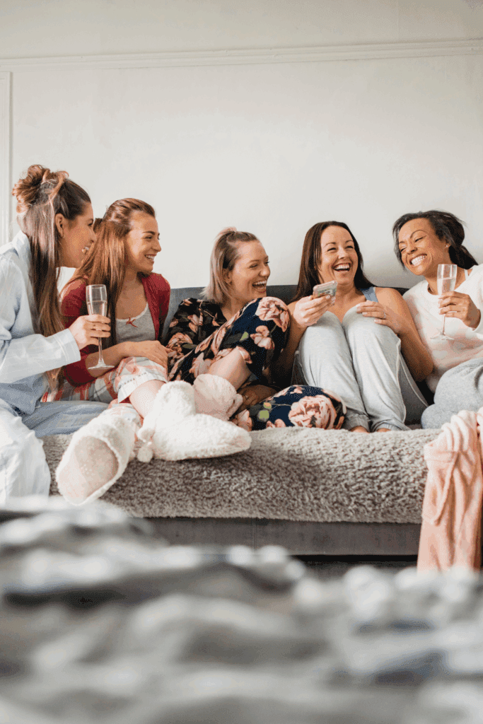 Group of five friends laughing together on a couch, enjoying drinks and sharing joyful moments in cozy loungewear.