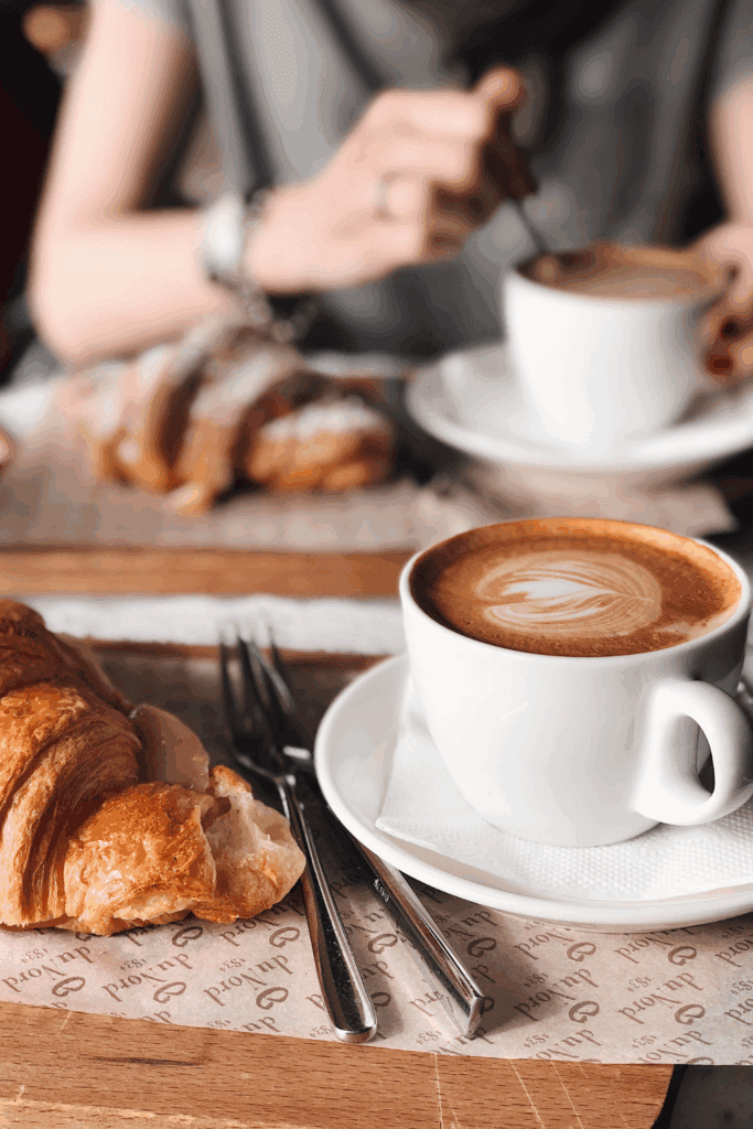 A close-up of a cozy cafe scene featuring a person stirring a cup of coffee and a croissant on a wooden table, with silverware and a napkin visible.