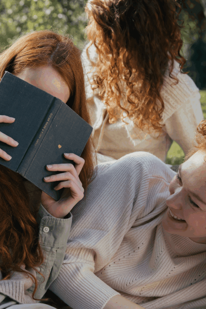 A group of friends enjoying a casual outdoor moment, with one friend playfully hiding behind a book while another smiles, creating a cozy and intimate atmosphere.