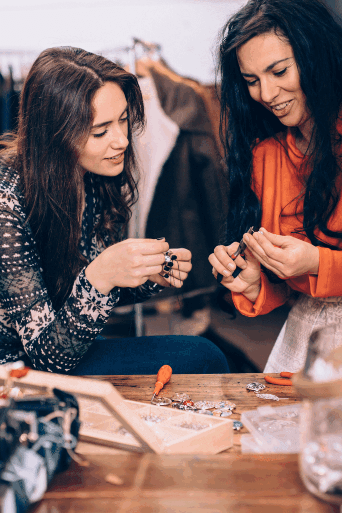 Two women engaged in a craft session, carefully working with jewelry-making materials at a wooden table, showcasing a creative and friendly atmosphere.