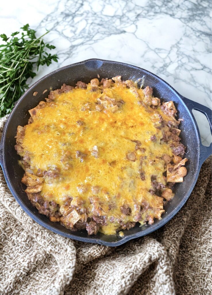 A skillet filled with a cheesy Sloppy Enchilada dish featuring ground beef, chickpeas, and tortilla pieces, topped with melted cheese, alongside fresh herbs and a cozy knit fabric.