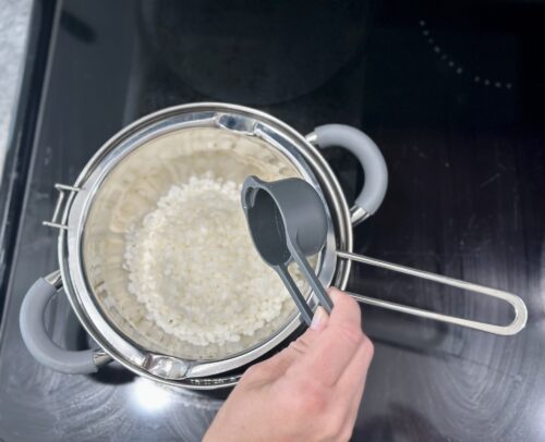 A person measuring beeswax pellets with a measuring cup over a double boiler setup on a stovetop.