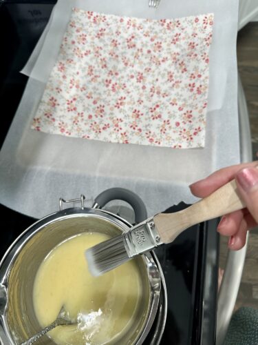 A hand holding a paintbrush over a pot of melted beeswax, with a floral-patterned cotton fabric placed on a sheet of parchment paper in the background.