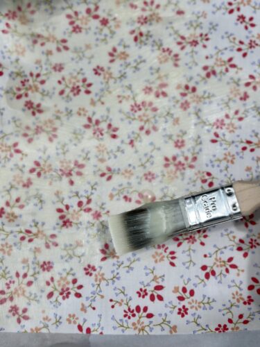 A paintbrush applying beeswax on a floral cotton fabric during the DIY beeswax wraps process.