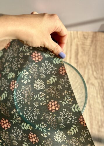 A hand holding a beeswax wrap over a glass bowl on a wooden surface, showcasing a floral pattern.