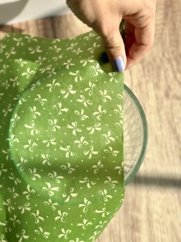 A hand is placing a green floral-patterned beeswax wrap over a glass bowl to cover it.