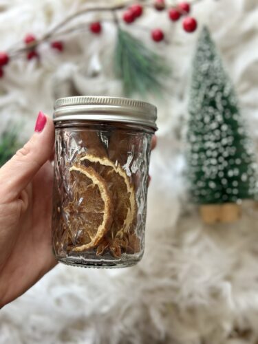 A person holding a glass jar filled with dried orange slices, surrounded by festive decorations including a small Christmas tree and red berries.