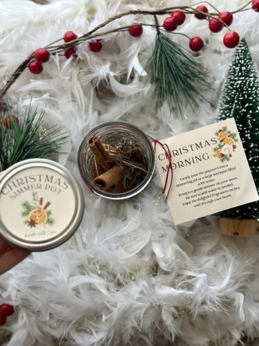 A small glass jar labeled 'Christmas Simmer Pot' contains dried orange slices and cinnamon sticks, with a tag that reads 'Christmas Morning' next to festive greenery and red berries.