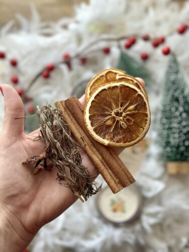 A hand holding a bundle of Christmas simmer pot ingredients, including dried orange slices, cinnamon sticks, rosemary sprigs, and star anise, with festive decorations in the background.
