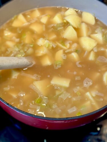 A close-up view of a pot filled with a savory soup containing diced potatoes, celery, and herbs, with a wooden spoon stirring the mixture.