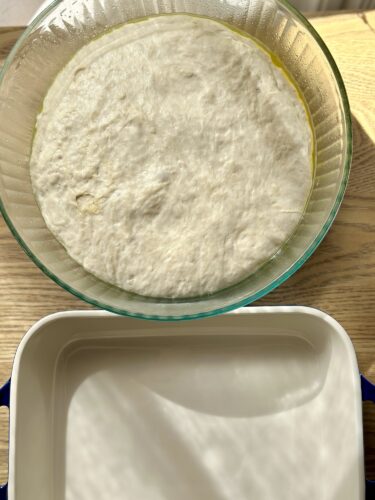 A bowl of dough rising for focaccia, with a preparing pan partially visible beneath it.