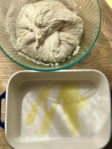 A bowl of risen focaccia dough sits above an oiled baking dish.