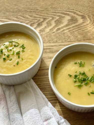 Two white bowls of vegan potato leek soup garnished with chopped chives, placed on a wooden table beside a linen towel.