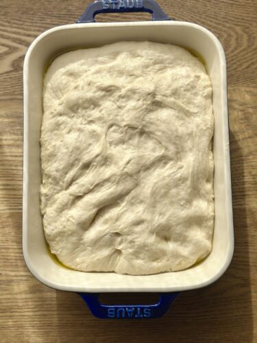 A pan of uncooked focaccia dough, spread out and resting before baking, with a glossy layer of olive oil visible around the edges.
