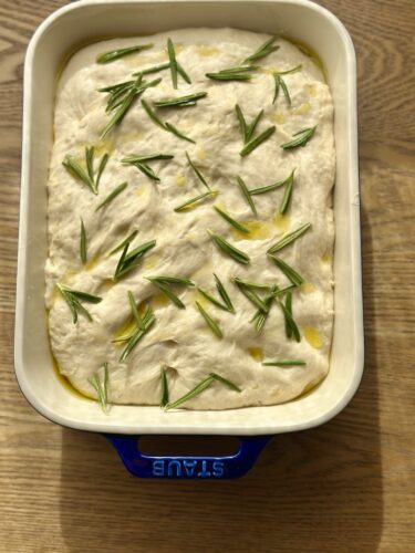 A baking dish filled with focaccia dough topped with fresh rosemary, drizzled with olive oil.