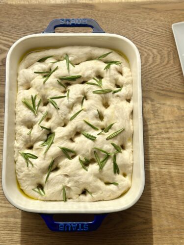 A pan of uncooked focaccia bread topped with fresh rosemary and drizzled with olive oil, resting on a wooden surface.
