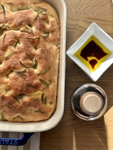 A freshly baked focaccia bread topped with rosemary, served in a ceramic dish, alongside a small dish of olive oil for dipping.