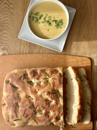 A freshly baked focaccia bread with a golden brown crust and rosemary sprigs, sliced and served next to a bowl of creamy soup topped with chives.