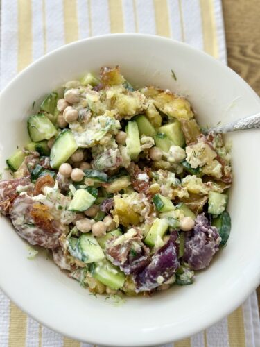 A creamy bowl of crispy smashed potato salad featuring chunky potatoes, diced cucumbers, and chickpeas, garnished with fresh herbs.