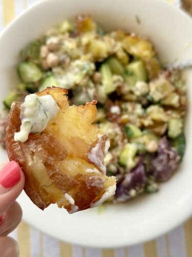 A crispy smashed potato being held, topped with tzatziki sauce, in front of a bowl of potato salad with cucumbers and chickpeas.