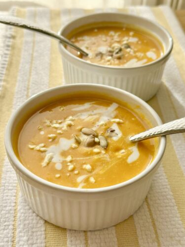 Close-up of two bowls of creamy pumpkin soup topped with a drizzle of cream and pumpkin seeds, placed on a textured kitchen towel.