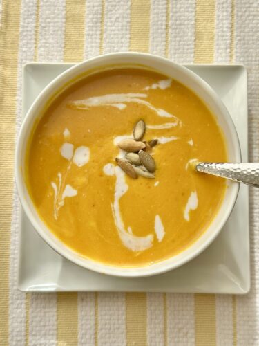 A bowl of creamy sweet potato and pumpkin soup topped with a swirl of coconut cream and pumpkin seeds, served on a white plate with a spoon.