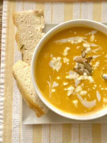 A bowl of creamy orange soup topped with seeds and drizzled with cream, served alongside slices of bread on a striped cloth.