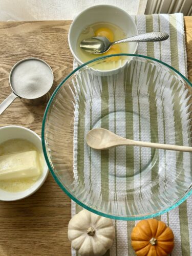 A glass mixing bowl with a wooden spoon, surrounded by bowls of beaten eggs, sugar, and melted butter, alongside small decorative pumpkins on a striped towel.