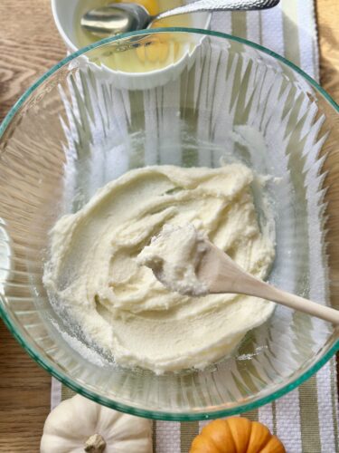 A mixing bowl with a creamy batter made from butter and sugar, a wooden spoon resting inside, with small decorative pumpkins and a striped towel in the background.