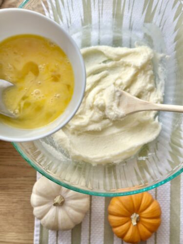 A bowl of beaten eggs next to a mixing bowl with creamy batter, with small white and orange pumpkins arranged nearby on a striped cloth.
