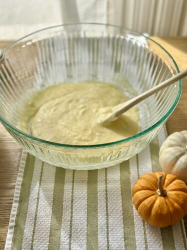 A mixing bowl filled with batter for pumpkin bread, resting on a striped kitchen towel, accompanied by small decorative pumpkins.