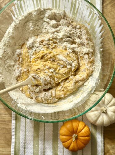 A glass mixing bowl containing a pumpkin bread batter being stirred, surrounded by flour, a small orange pumpkin, and a white pumpkin on a kitchen towel.