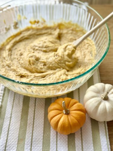 A glass bowl filled with pumpkin bread batter, with a wooden spoon resting inside, alongside small decorative pumpkins on a striped kitchen towel.