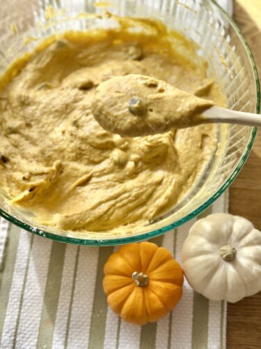 A bowl of pumpkin bread batter with a wooden spoon, flanked by small decorative pumpkins on a striped cloth.