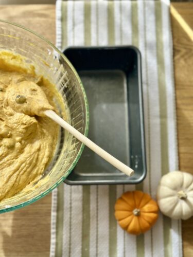 A bowl of pumpkin bread batter with a wooden spoon, next to an empty loaf pan and small decorative pumpkins on a striped kitchen towel.