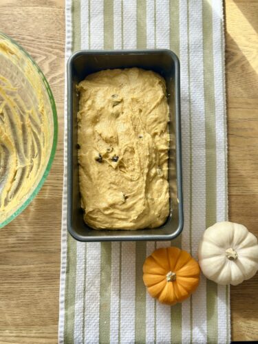 A loaf pan filled with unbaked pumpkin bread batter sits on a striped kitchen towel, accompanied by two small pumpkins—one orange and one white.