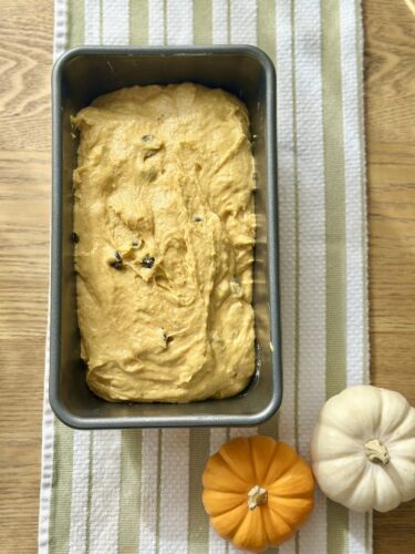 A loaf pan filled with pumpkin bread batter, topped with chocolate chips, placed on a striped kitchen towel alongside small pumpkins.