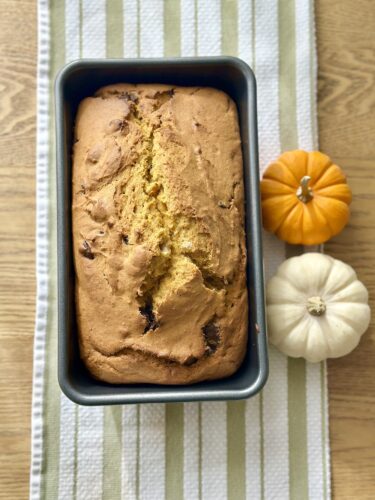 A freshly baked loaf of pumpkin bread in a grey loaf pan, surrounded by small decorative pumpkins on a striped linen cloth.