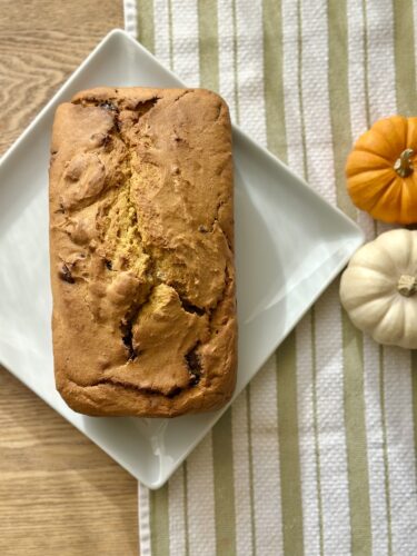 A golden loaf of pumpkin bread cooling on a white plate, surrounded by small decorative pumpkins, with a striped kitchen towel in the background.