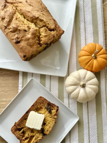 A loaf of pumpkin bread topped with a pat of butter, placed on a white plate, with additional mini pumpkins in orange and white surrounding it, all set against a striped table runner.