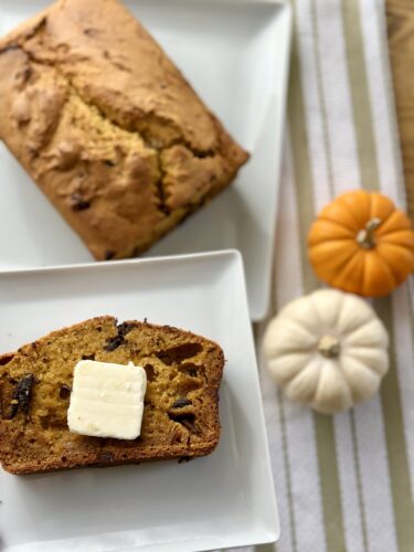 A slice of freshly baked pumpkin bread topped with a pat of butter, placed on a white plate beside the whole loaf, with small decorative pumpkins in the background.