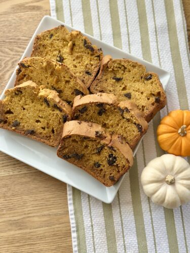 Sliced pumpkin bread with chocolate chips arranged on a square plate, accompanied by small decorative pumpkins, on a striped towel.